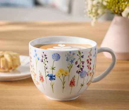 Kvetinový hrnček s kávou a latte art srdce na drevenom stole vedľa taniera s pečivom a vázy s kvetmi.