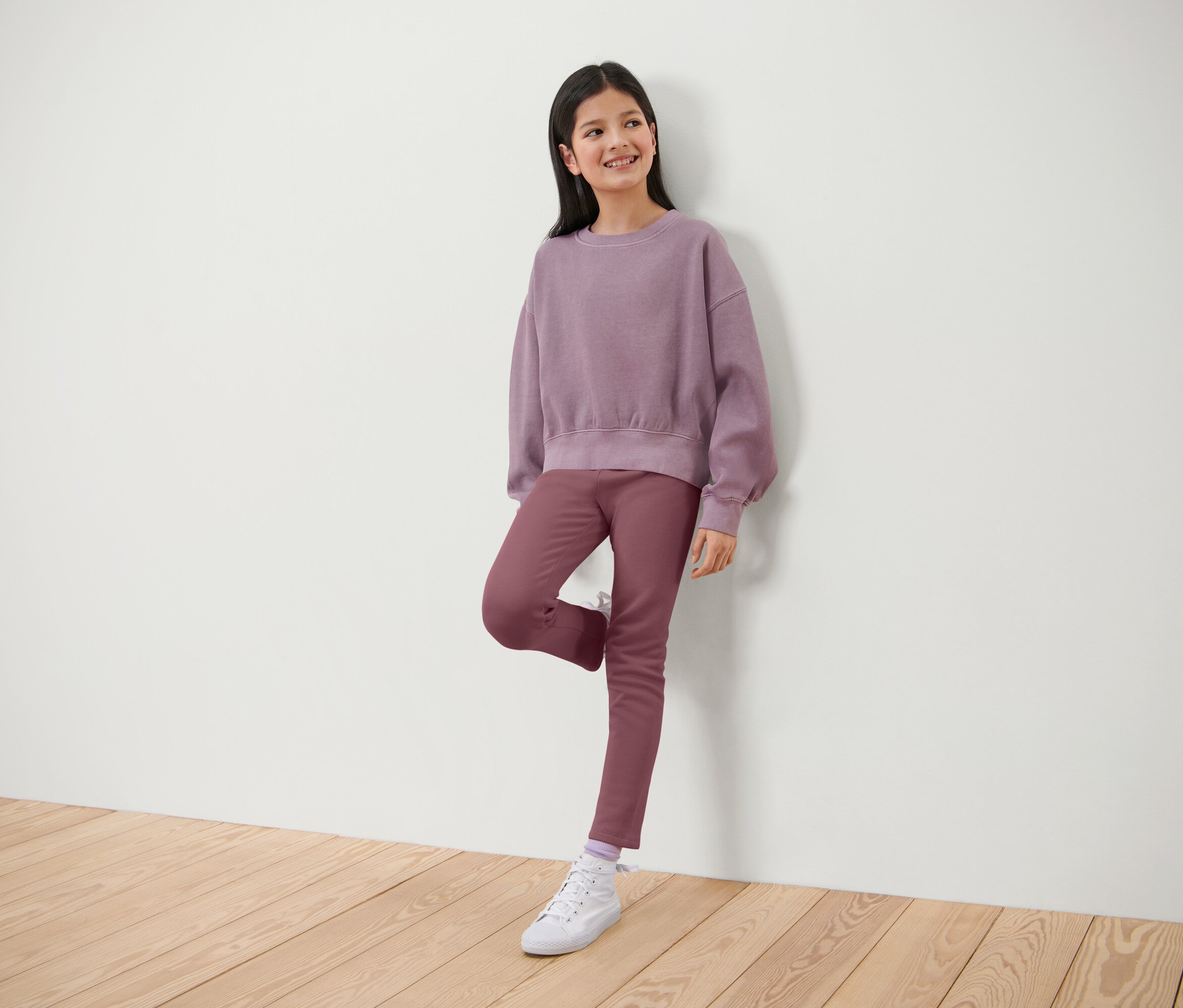 Usmievajúce sa dievča pózuje v lila svetri a nohaviciach, opiera sa o bielu stenu.