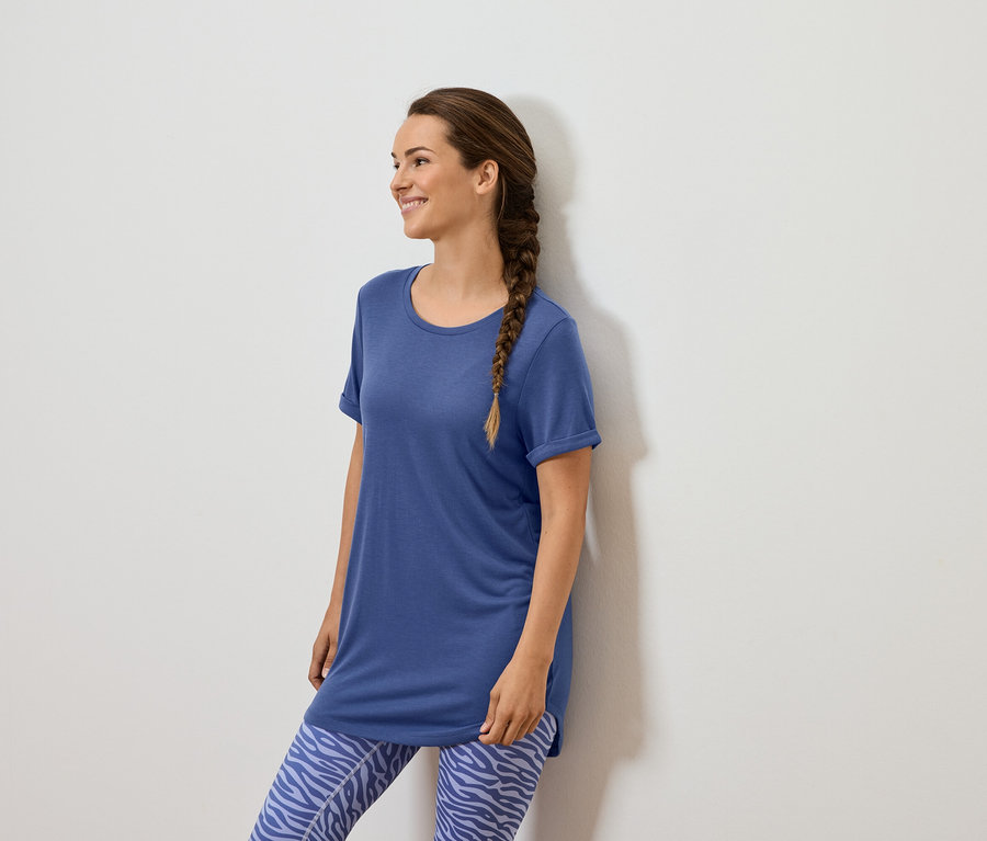 Žena s hnedým vrkočom stojí pri bielej stene a má na sebe modré tričko a modré legíny so zvieracím vzorom.