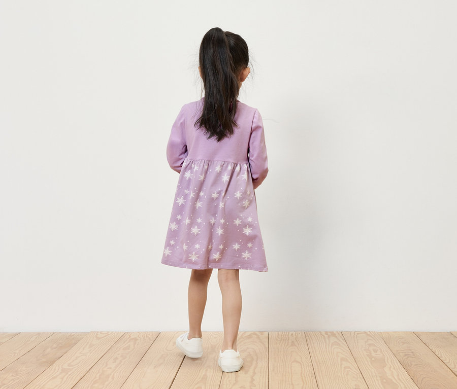 Dievča v lila šatách s hviezdami a bielych teniskách stojí chrbtom k fotoaparátu.