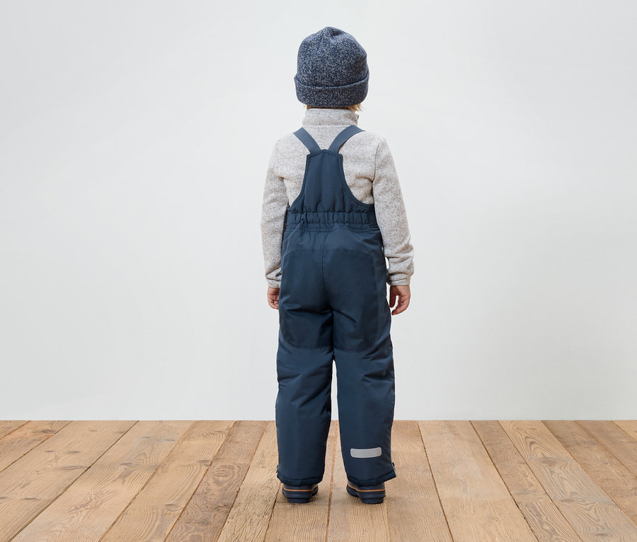 Chlapec chrbtom k fotoaparátu má na sebe modré nohavice s náprsenkou, sivý sveter a modrú čiapku.