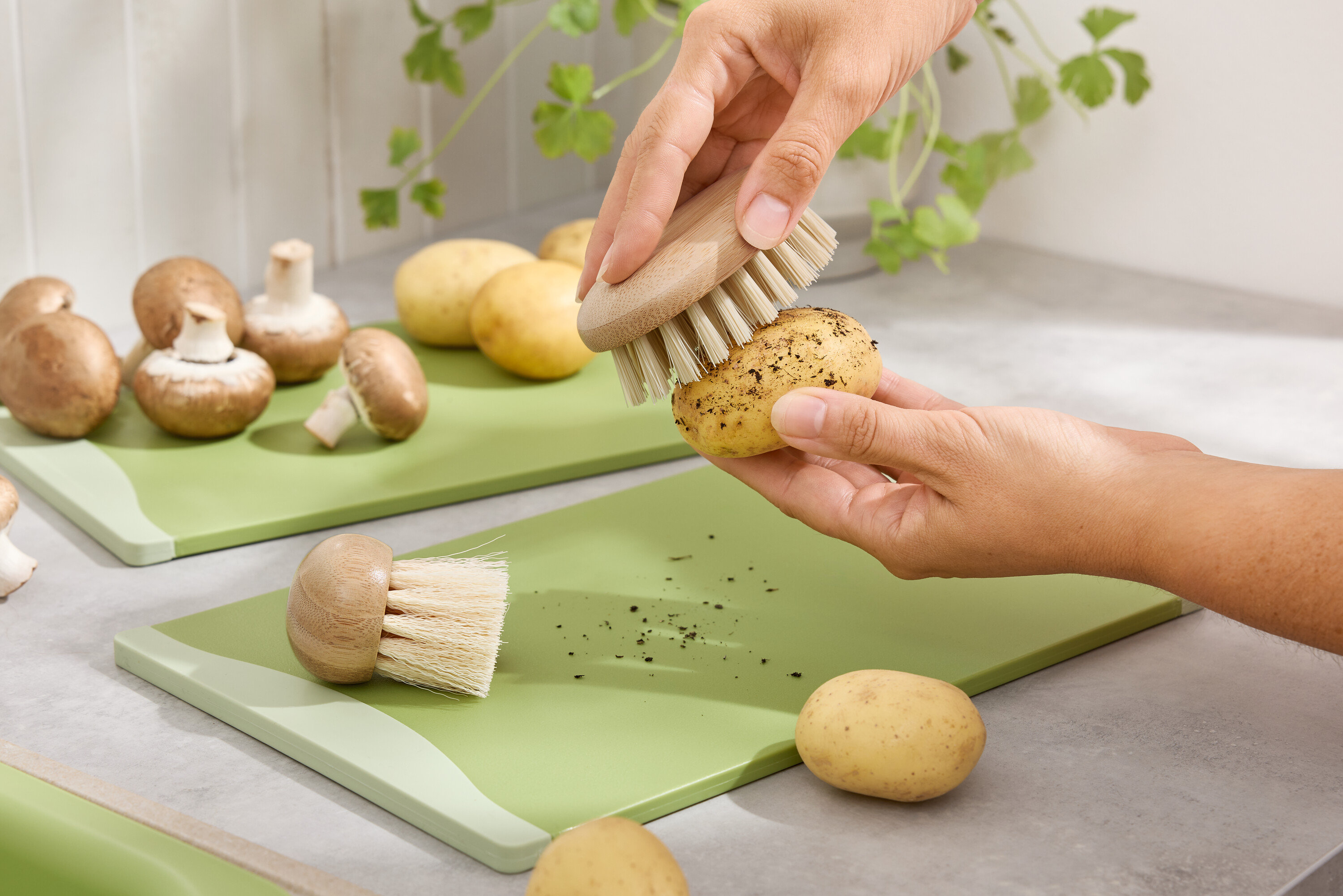 Ruky čistia kefou zemiak na zelenej doske na krájanie. Hnedé šampiňóny a zemiaky sú v pozadí.