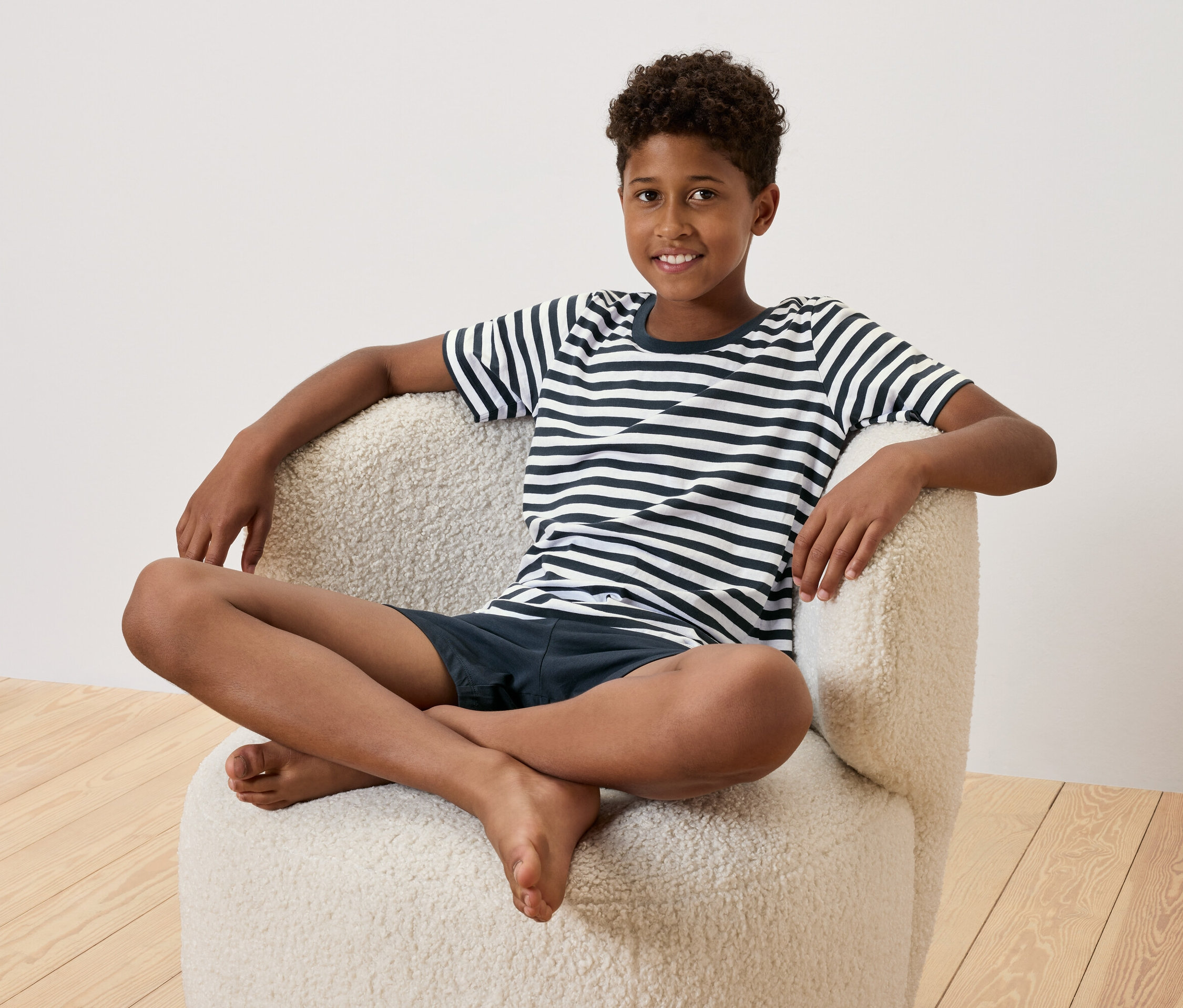 Mladý chlapec sedí so skríženými nohami v krémovom kresle. Má na sebe pruhované tričko a tmavomodré šortky.