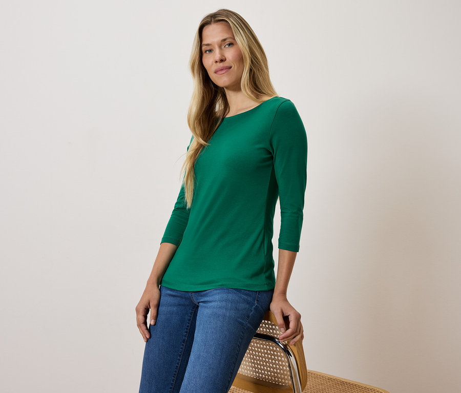 Blondínka v zelenom tričku s trojštvrťovým rukávom a modrých džínsoch sedí na stoličke.