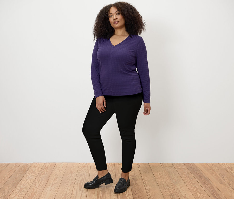 Žena pózuje v štruktúrovanom tričku s dlhým rukávom slivkovej farby a v džegínsoch.