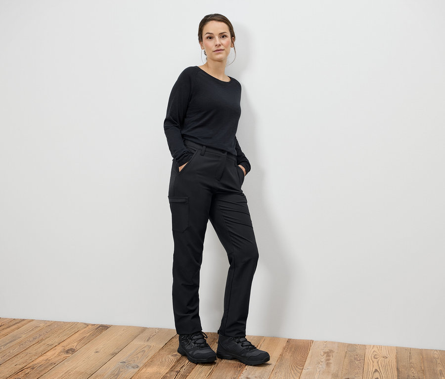 Žena stojí s rukami vo vreckách v čiernych nohaviciach a čiernych topánkach.