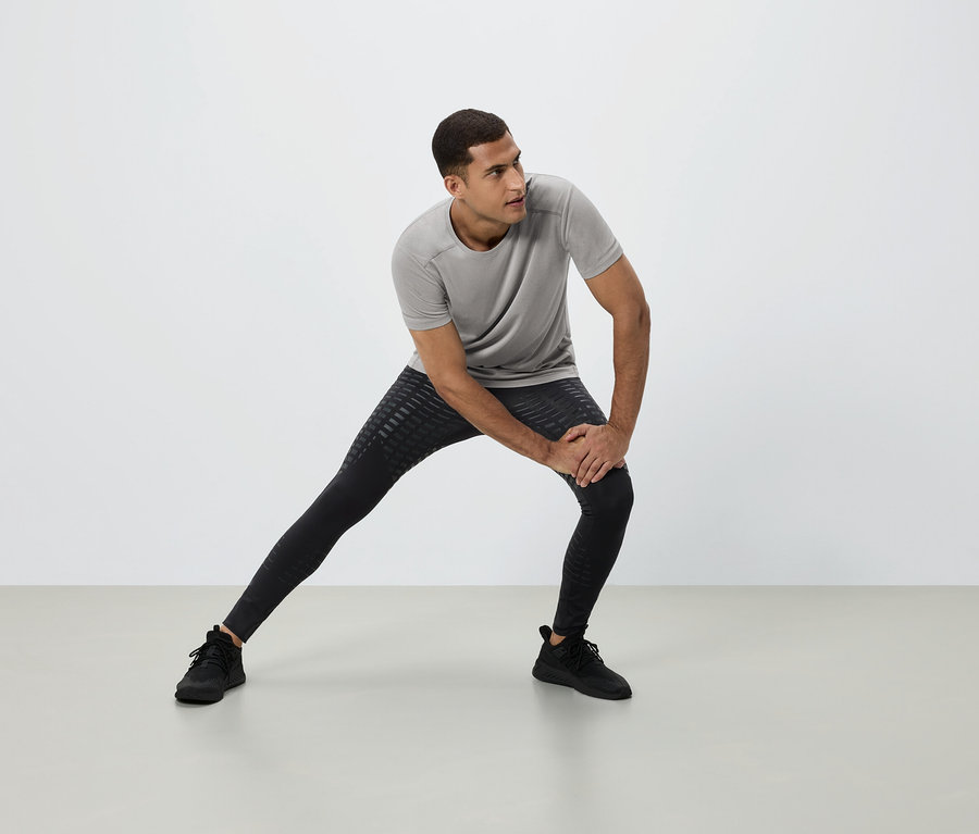 Muž sa naťahuje v sivom tričku, čiernych legínach a čiernych teniskách na bielom pozadí.