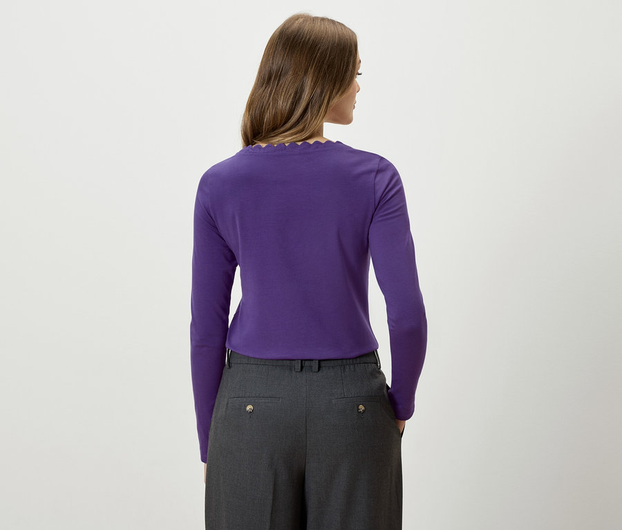 Žena chrbtom k fotoaparátu má na sebe fialové tričko s dlhým rukávom a sivé nohavice.