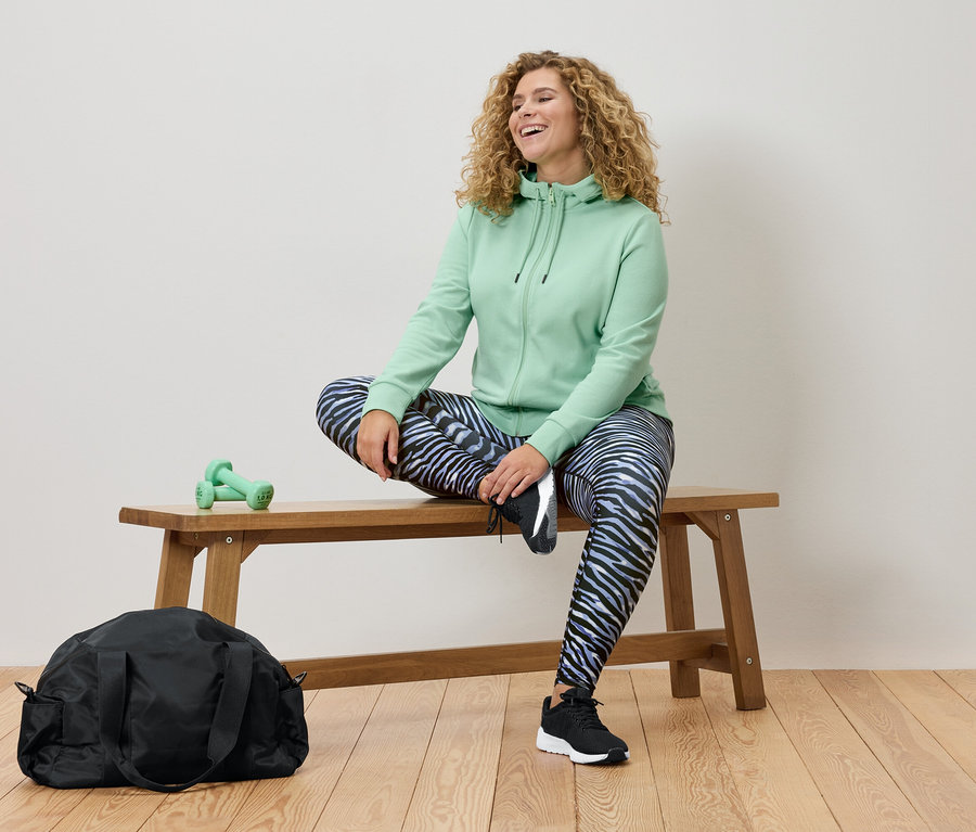 Žena s kučeravými vlasmi sedí na drevenej lavičke, má na sebe mätovú mikinu s kapucňou a legíny so vzorom zebry. Viditeľná je čierna športová taška a zelené činky.