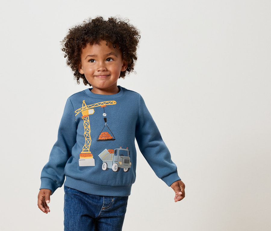 Chlapec s kučeravými vlasmi má na sebe modrú mikinu s aplikáciou žltého žeriavu a nákladného auta.