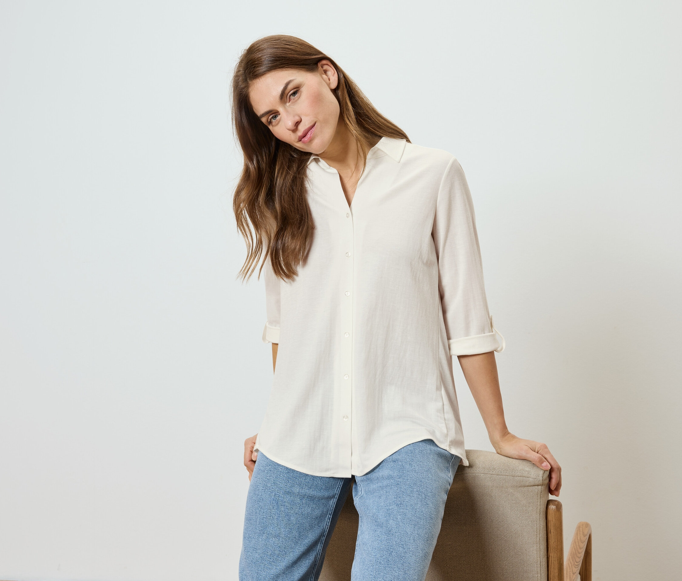 Žena s vlnitými vlasmi, oblečená do svetlej košele a džínsov, opiera sa o stoličku.