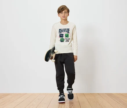 Chlapec drží skateboard a má na sebe svetlé tričko s dlhým rukávom s motívom Minecraftu, kombinované s tmavými teplákmi a teniskami na jednoduchom pozadí.