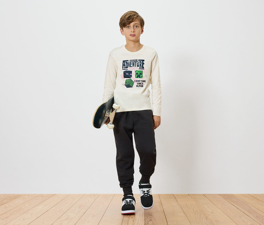 Chlapec drží skateboard a má na sebe svetlé tričko s dlhým rukávom s motívom Minecraftu, kombinované s tmavými teplákmi a teniskami na jednoduchom pozadí.