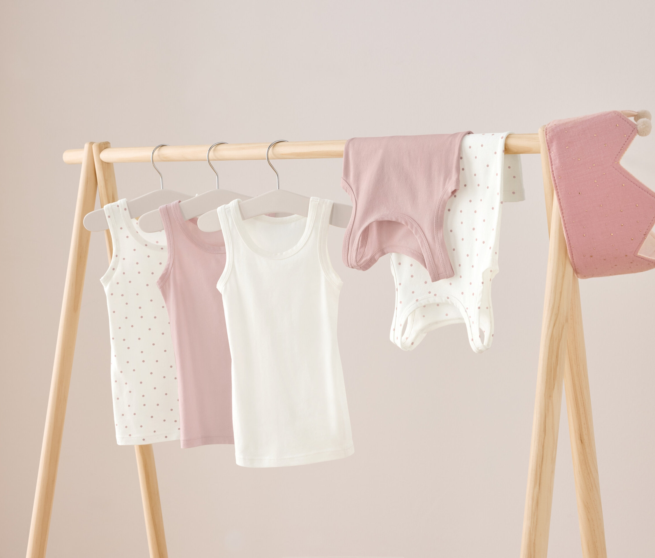 Na drevenom stojane visia tri tielka v bielej, ružovej a bielej farbe s bodkami, ako aj ružová košeľa a biele body s bodkami. Vpravo visí ružová koruna.