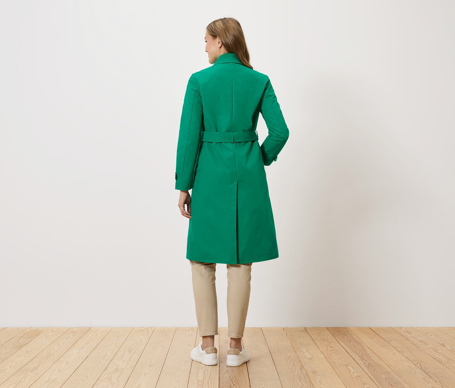 Žena stojí chrbtom k fotoaparátu a má na sebe zelený kabát s opaskom, béžové nohavice a biele tenisky.