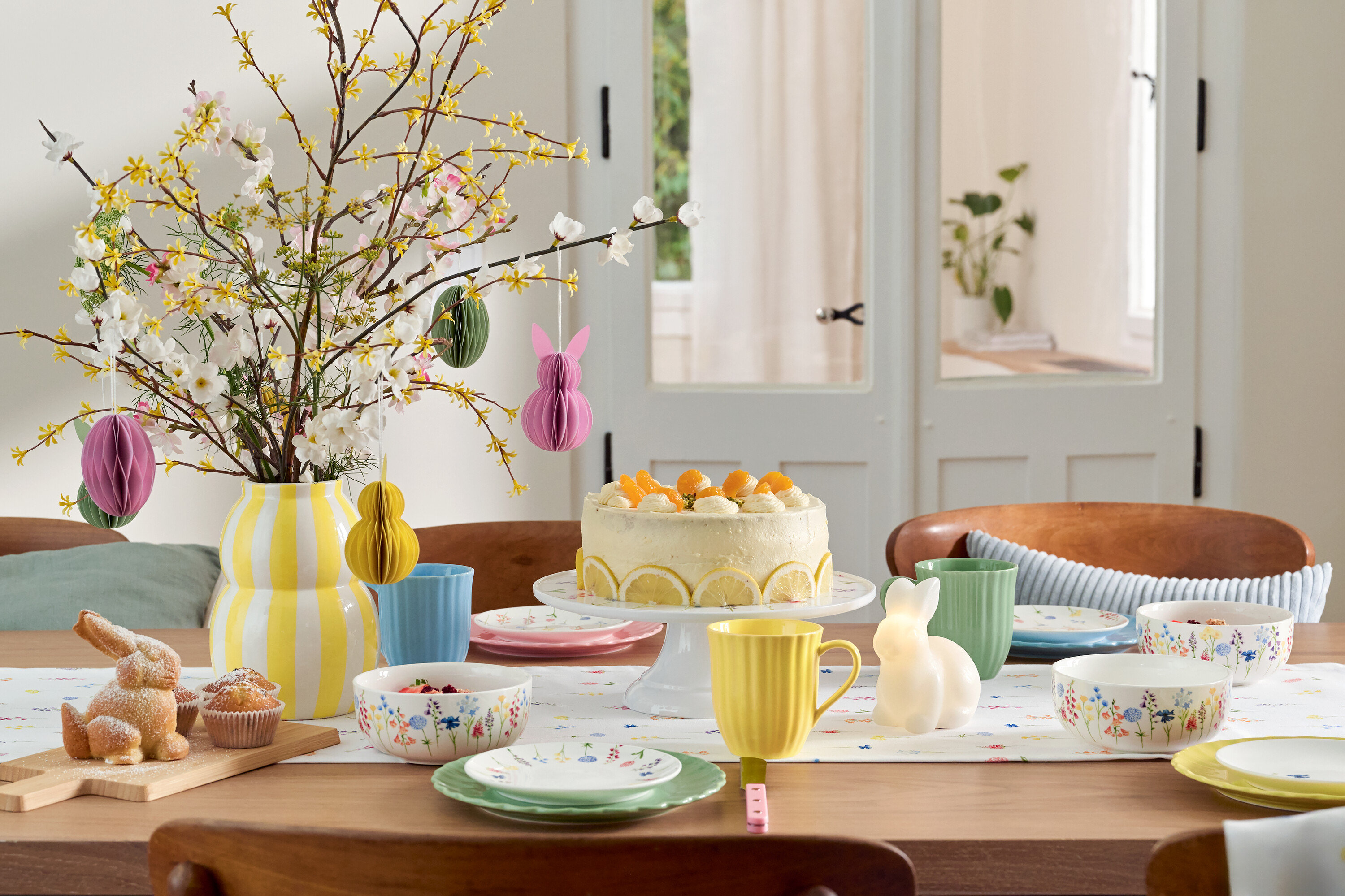 Jedálenský stôl s veľkonočnou výzdobou, koláčom, riadom a vázou s kvetmi.