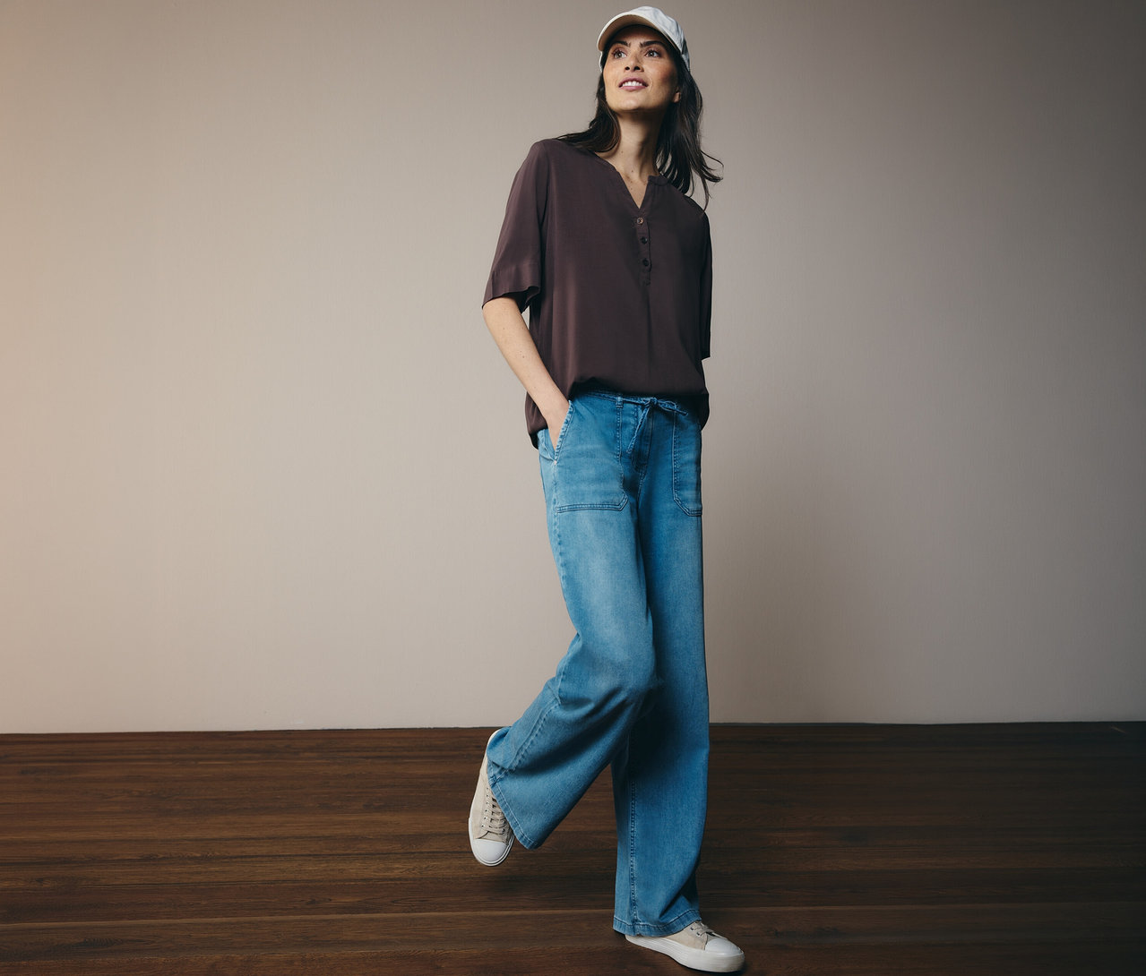 Žena v šiltovke a hnedej tunikovej blúzke CECIL, džínsoch a teniskách.