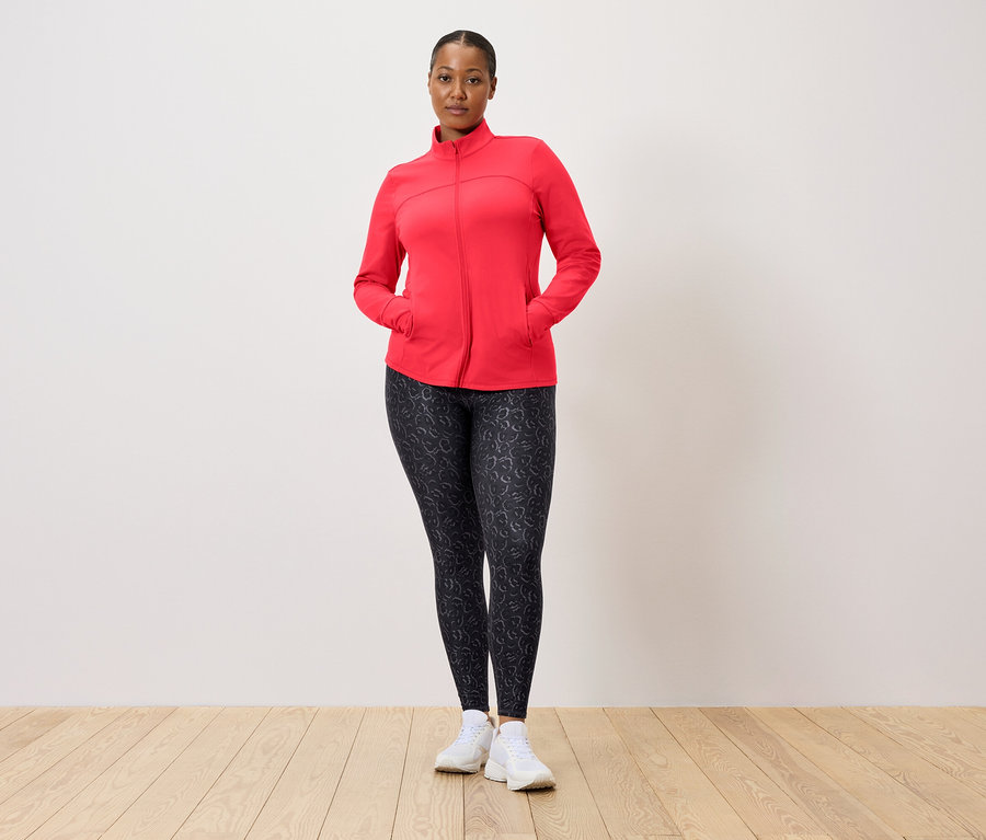 Žena v červenej bunde a vzorovaných legínach pózuje pred stenou.