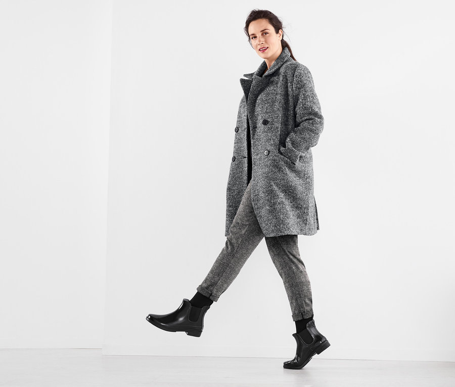 Žena pózuje v šedom kabáte, tričku s vodopádovým výstrihom, nohaviciach a termopodkolienkach.