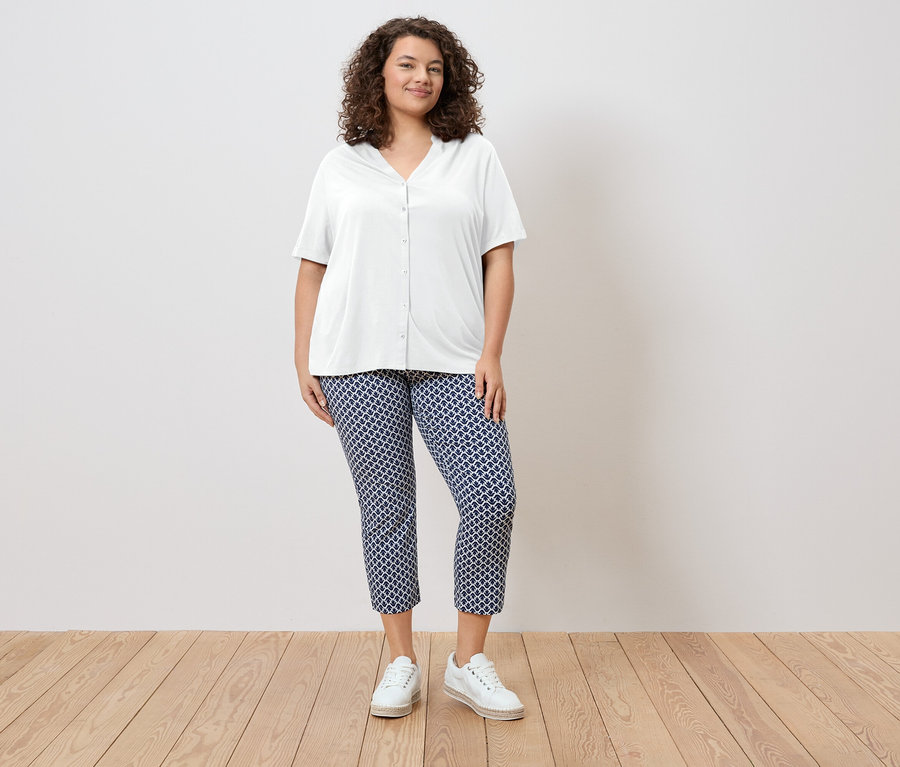 Žena s kučeravými vlasmi má na sebe bielu blúzku a modré vzorované nohavice a biele topánky.