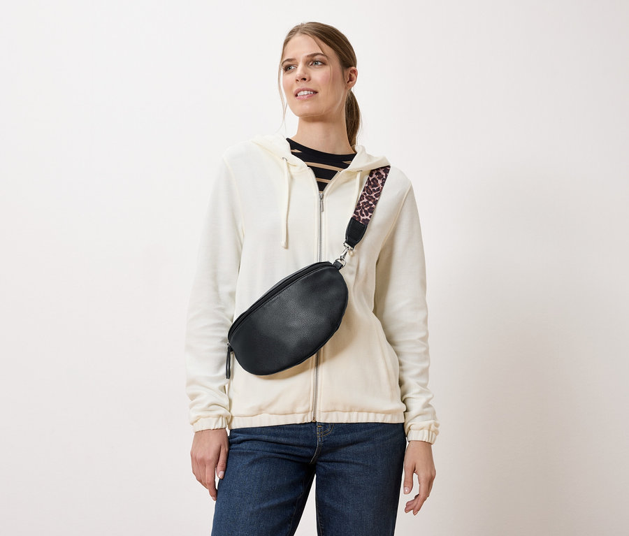 Žena nosí čiernu bedrovú tašku s popruhom s leopardím vzorom cez krémovú bundu a džínsy.