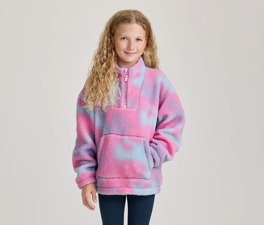 Usmievavé dievča s kučeravými blond vlasmi v ružovo-fialovo-modrom tie-dye flísovom pulóvri s polovičným zipsom a vreckom.