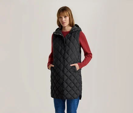 Žena v dlhej čiernej prešívanej veste s kapucňou, červenom tričku s dlhým rukávom a modrých džínsoch, s rukami vo vreckách.
