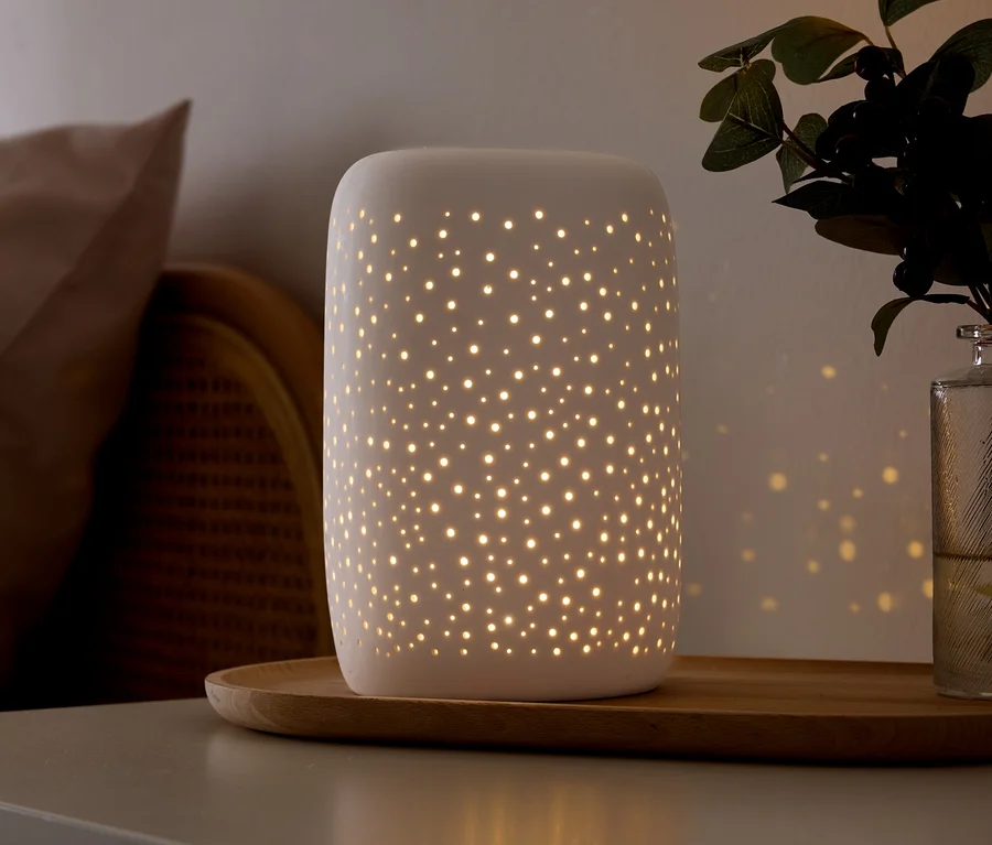 Biela keramická lampa s malými perforáciami vyžaruje teplé svetlo a vrhá hviezdne vzory na stenu. Stojí na drevenom podnose na sivom povrchu, vedľa béžového vankúša, prúteného nábytku a zelenej rastliny v sklenenej váze.