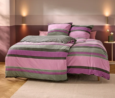 Posteľ s pruhovanou posteľnou bielizňou v ružovej, fialovej a olivovozelenej farbe. Dva zladené vankúše a jeden malý ružový vankúš. Steny sú dvojfarebné v svetloružovej a tmavoružovej/fialovej, s dvoma nástennými svietidlami. Napravo je odkladací stolík s vázou bielych kvetov. Drevená podlaha s béžovým kobercom.