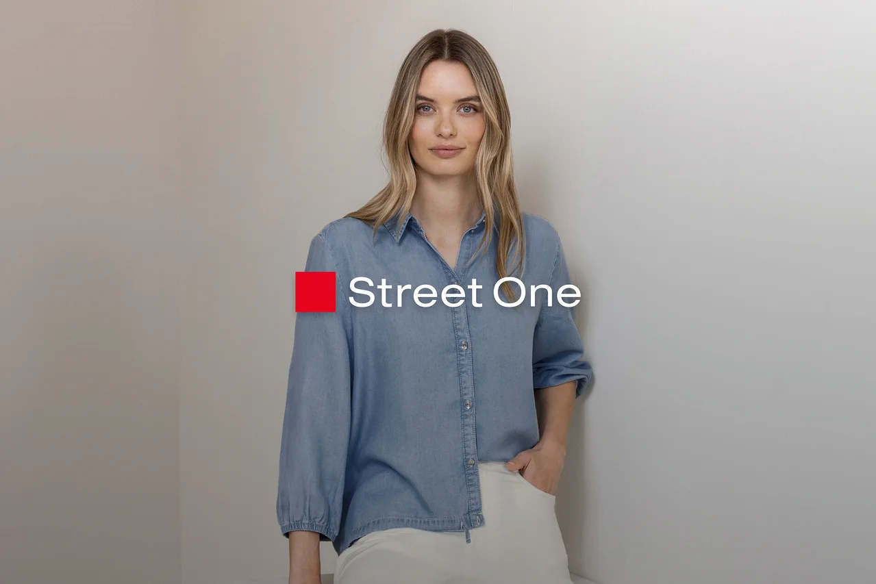 Blondínka v svetlomodrej džínsovej košeli a svetlých nohaviciach, stojí s rukou vo vrecku pri sivej stene. Logo Street One.