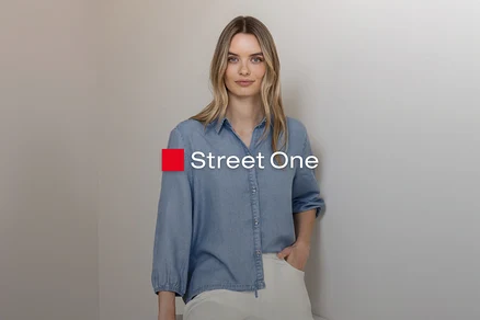 Blondínka v svetlomodrej džínsovej košeli a svetlých nohaviciach, stojí s rukou vo vrecku pri sivej stene. Logo Street One.
