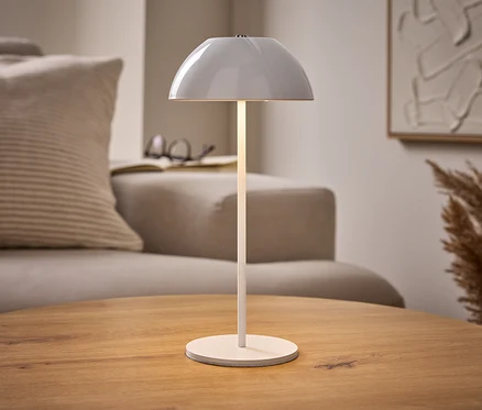 Osvetlená biela stolová LED lampa s akumulátorom na drevenom stole, s béžovou pohovkou, knihou a abstraktným umením v pozadí.
