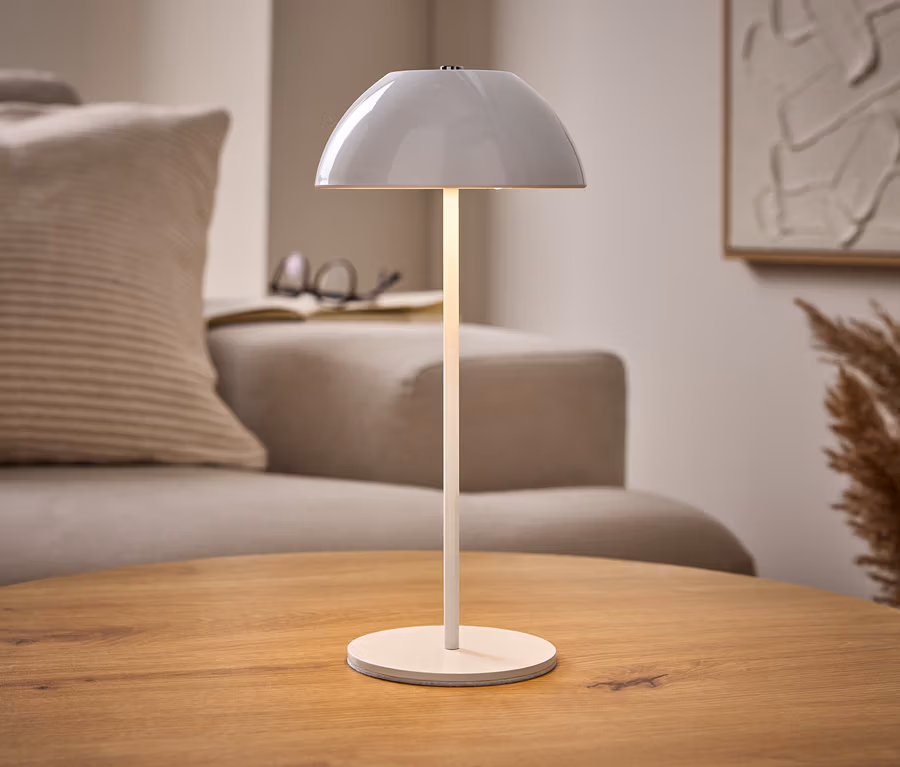 Osvetlená biela stolová LED lampa s akumulátorom na drevenom stole, s béžovou pohovkou, knihou a abstraktným umením v pozadí.
