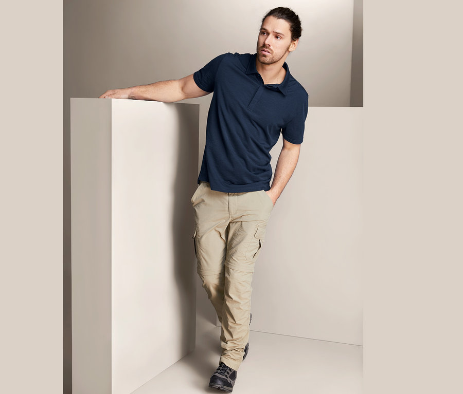 Muž opretý o bielu stenu má na sebe tmavomodré funkčné polo tričko s merino vlnou a béžové nohavice.