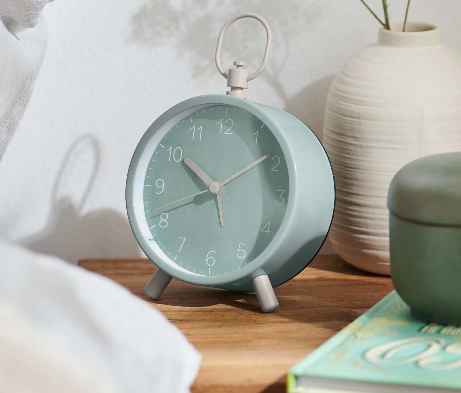 Mätovozelený analógový dizajnový budík stojí na drevenom nočnom stolíku.