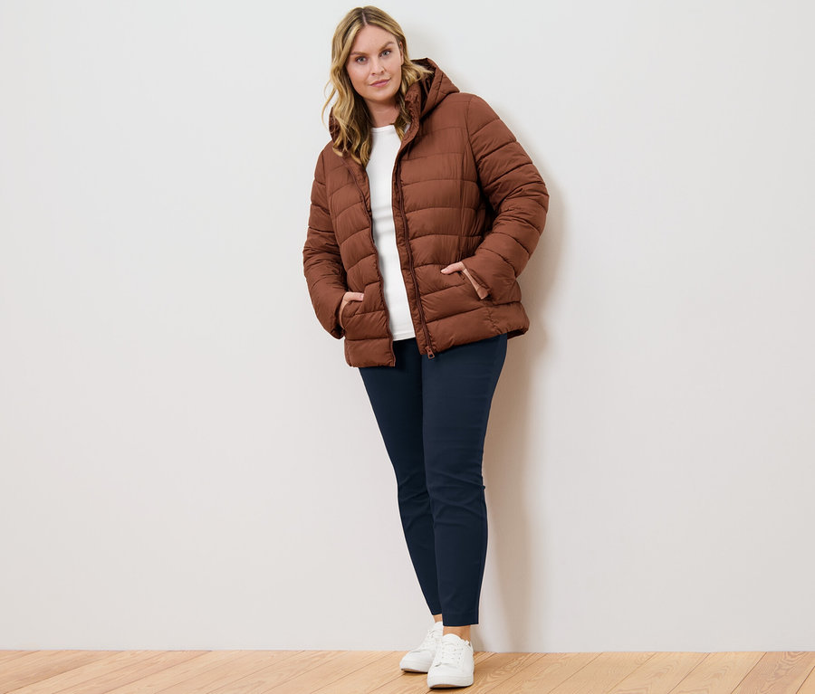 Žena pózuje v koňakovej prešívanej bunde s kapucňou, tmavomodrých elastických nohaviciach a bielom tričku s trojštvrťovými rukávmi.