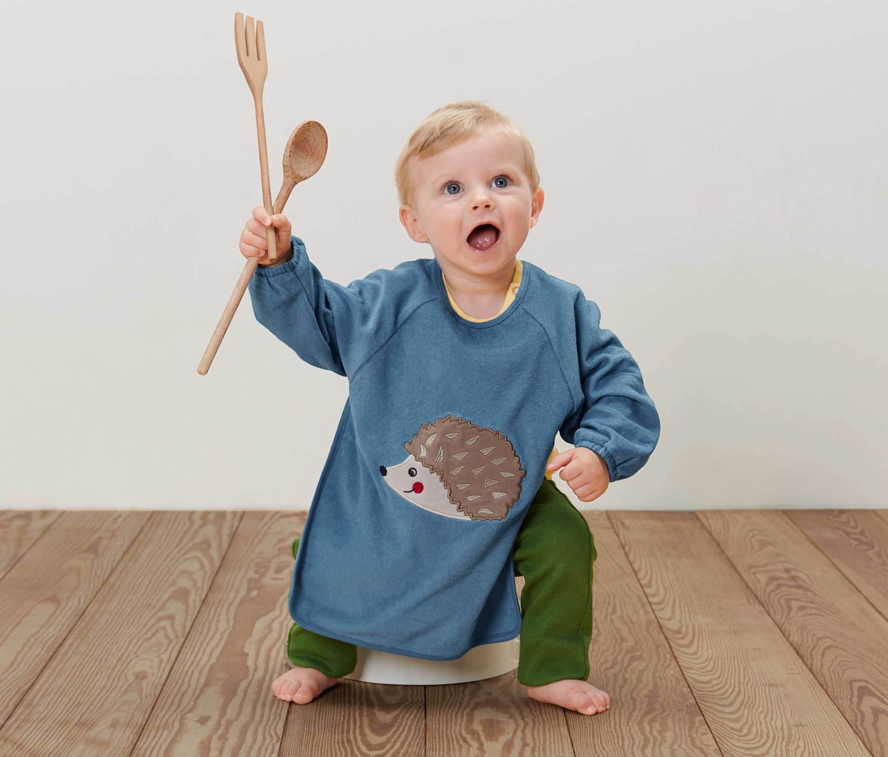 Batoľa sedí na zemi a má na sebe modrý detský podbradník s dlhým rukávom s ježkom a drží drevenú lyžicu a vidličku.