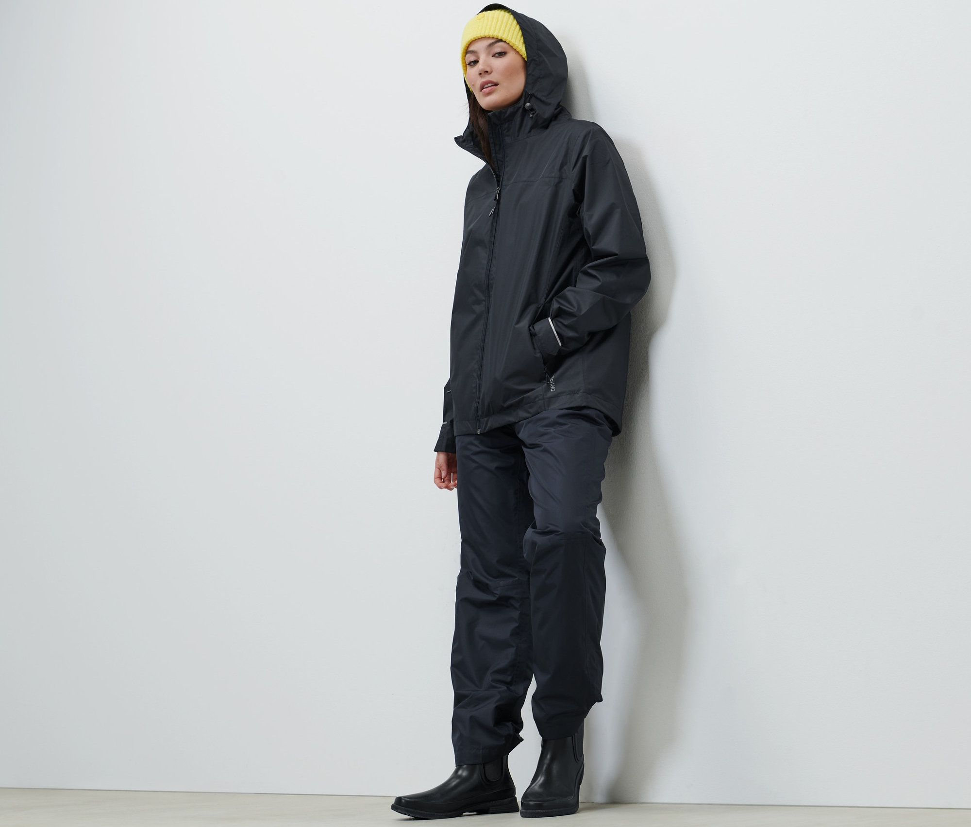 Žena má na sebe tmavomodrú unisexovú bundu do dažďa, nohavice a žltú čiapku.