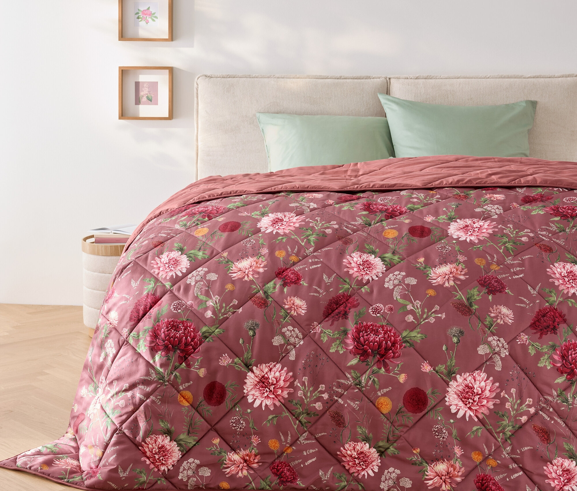 Na posteli leží prešívaná prikrývka s kvetinovým vzorom a dva zelené vankúše.