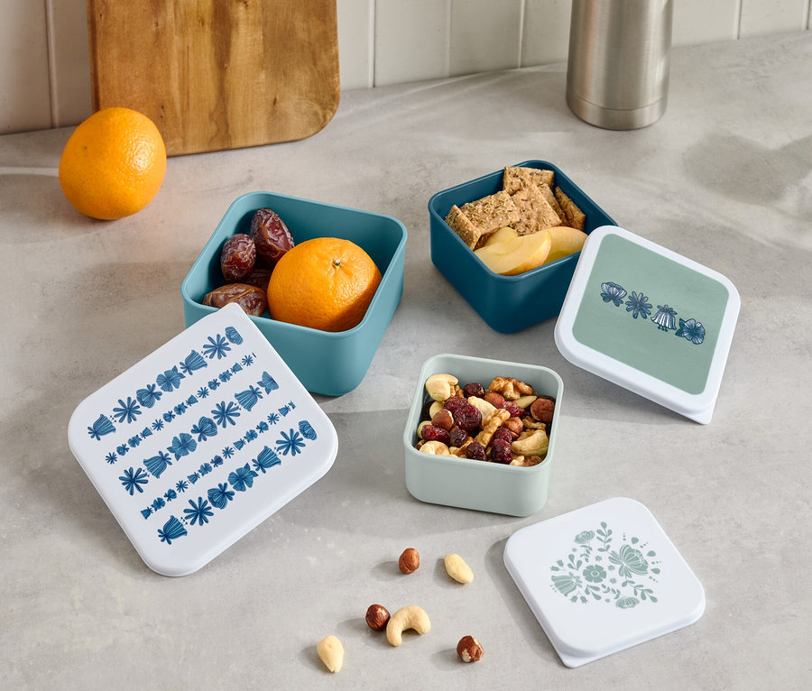 Na linke stojí termoska a drevená doska na krájanie vedľa štyroch malých otvorených krabičiek s ovocím, krekrami a orechmi a štyroch viečok s rôznymi vzormi.