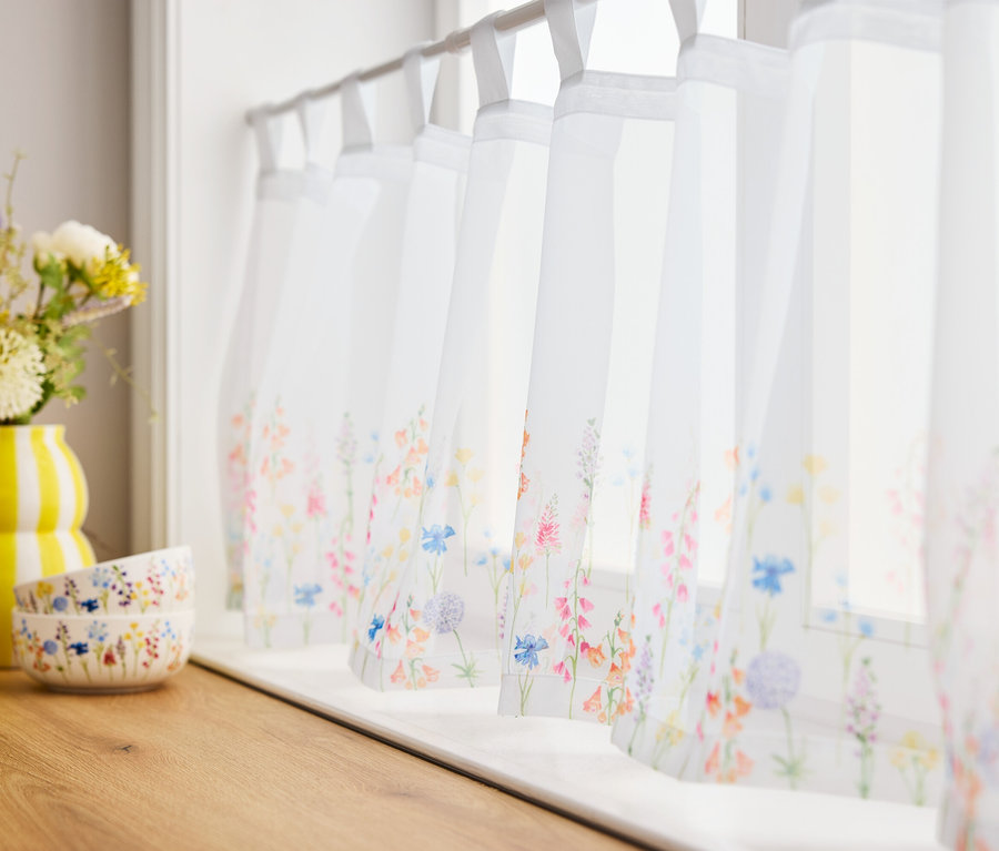 Kvety v žltej váze a dve misky na drevenej doske pred oknami so záclonami s kvetinovým vzorom.