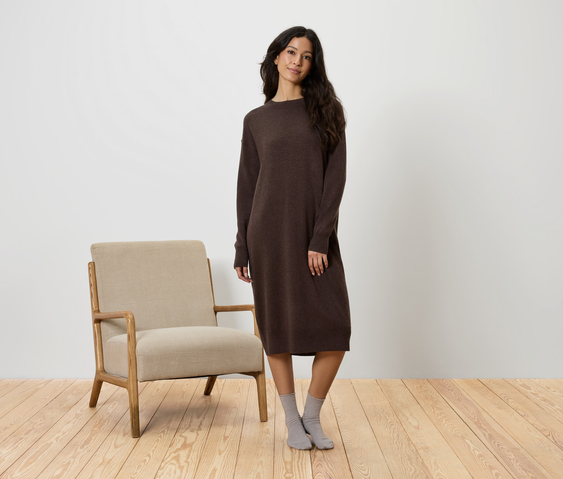Žena stojí vedľa béžového kresla a má na sebe hnedé šaty a sivé ponožky.