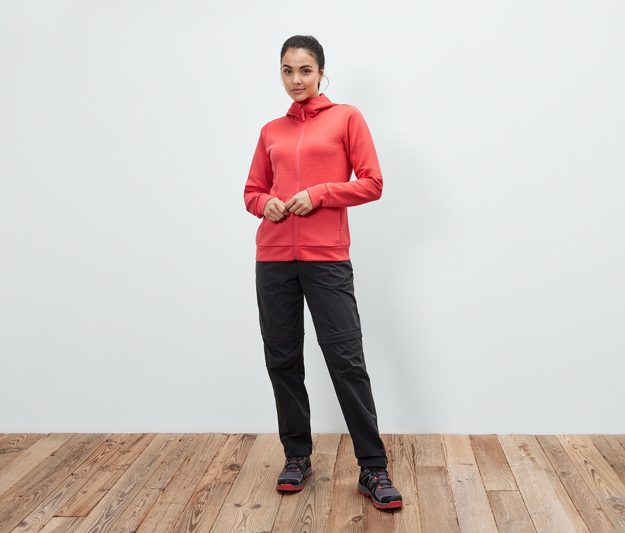 Žena pózuje v koralovej bunde s kapucňou, čiernych nohaviciach a topánkach na drevenej podlahe.