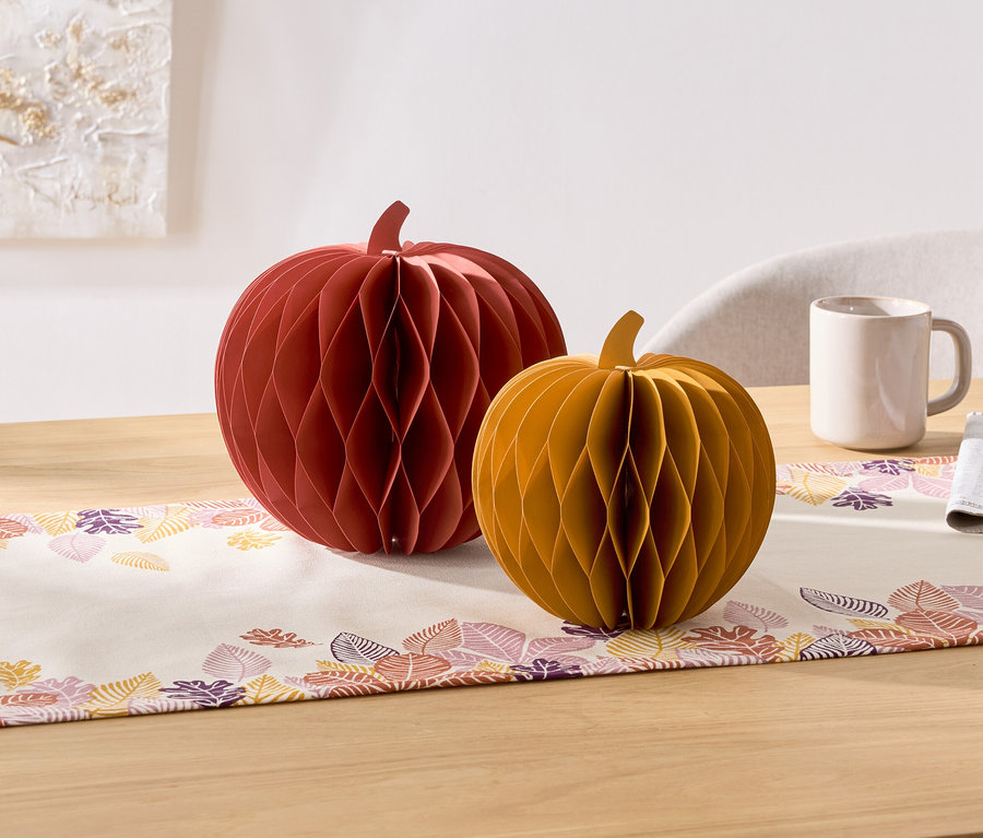 Na stole sú dve dekoratívne papierové tekvice, červená a žltá. Vedľa nich je hrnček a servítka.