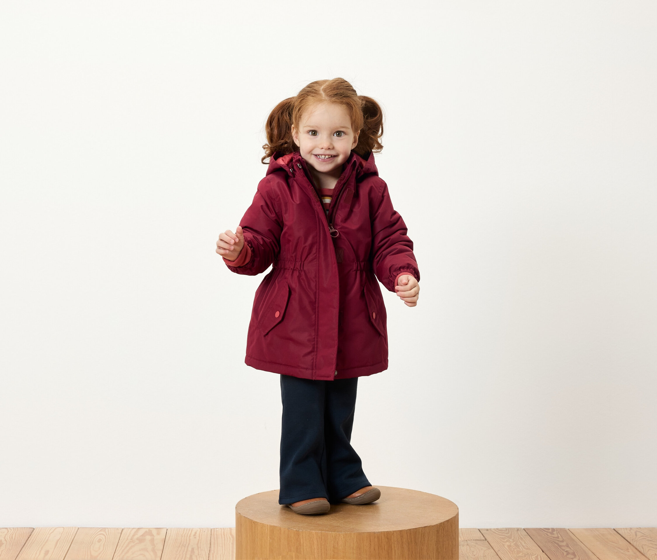 Dievča s ryšavými vrkočmi stojí na drevenom podstavci v bordovej bunde a tmavomodrých nohaviciach.