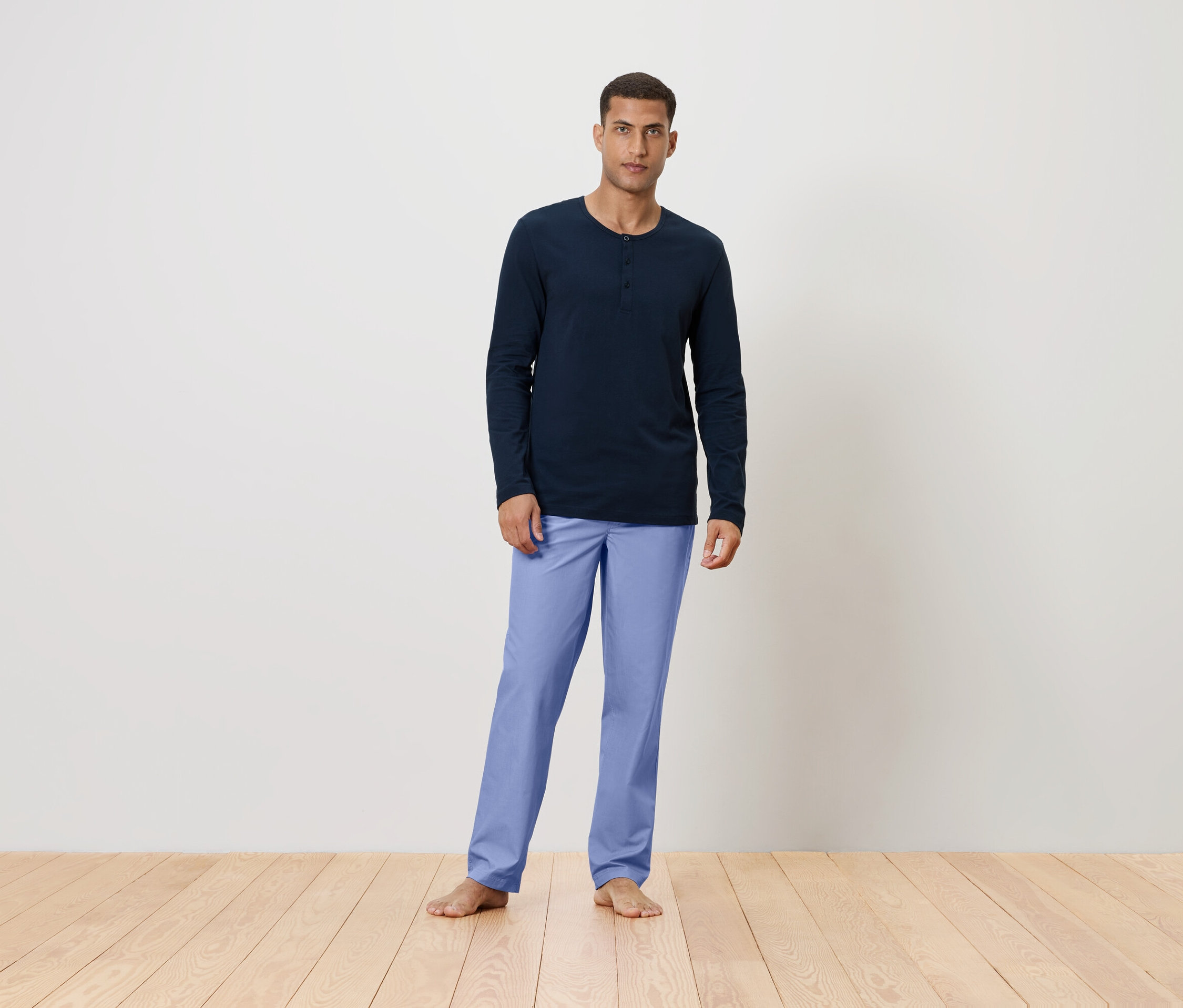 Muž stojí bosý na drevenej podlahe, má na sebe tmavomodré tričko s dlhým rukávom a svetlomodré nohavice.
