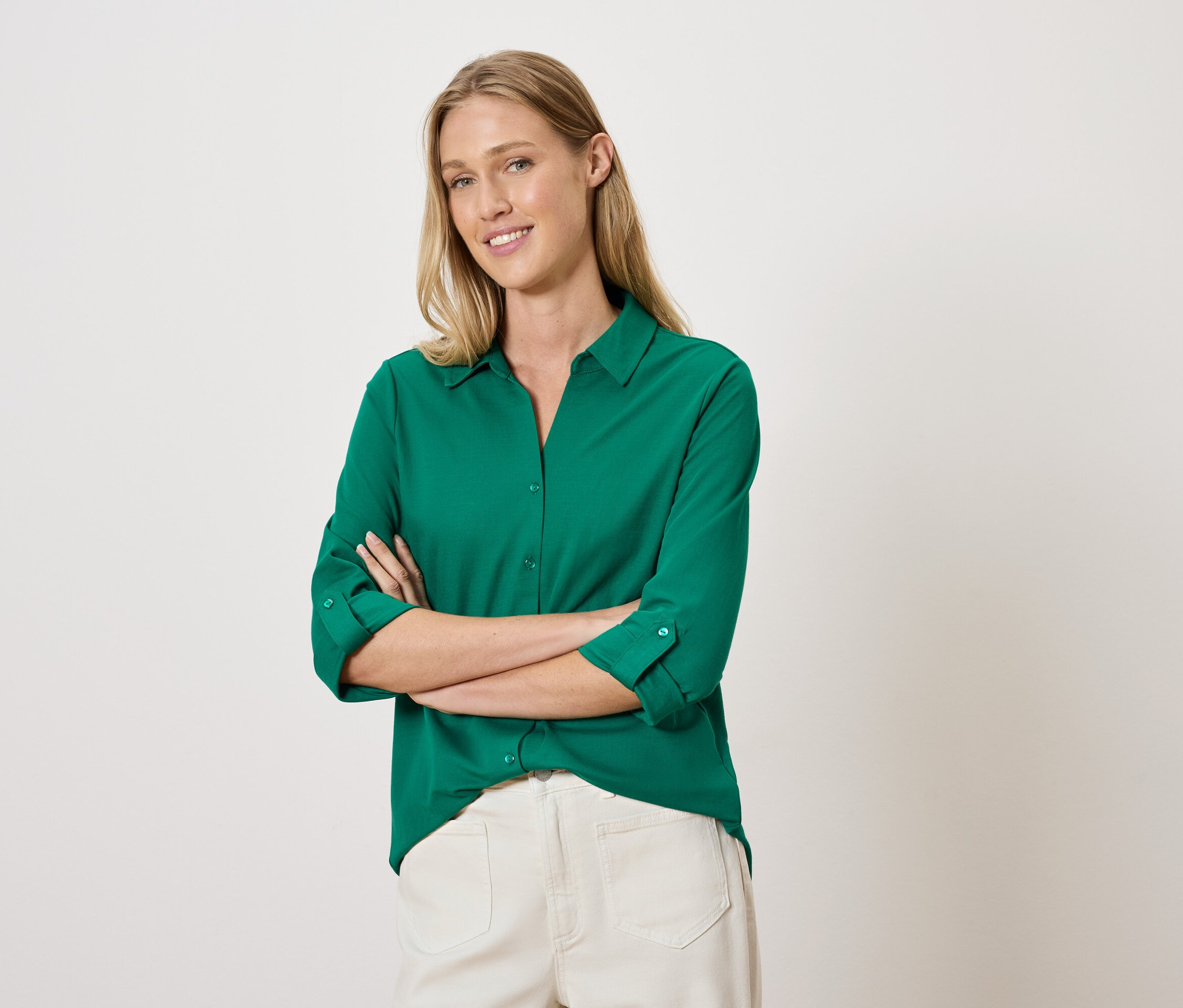 Žena pózuje so skríženými rukami v zelenej košeli a bielych nohaviciach.