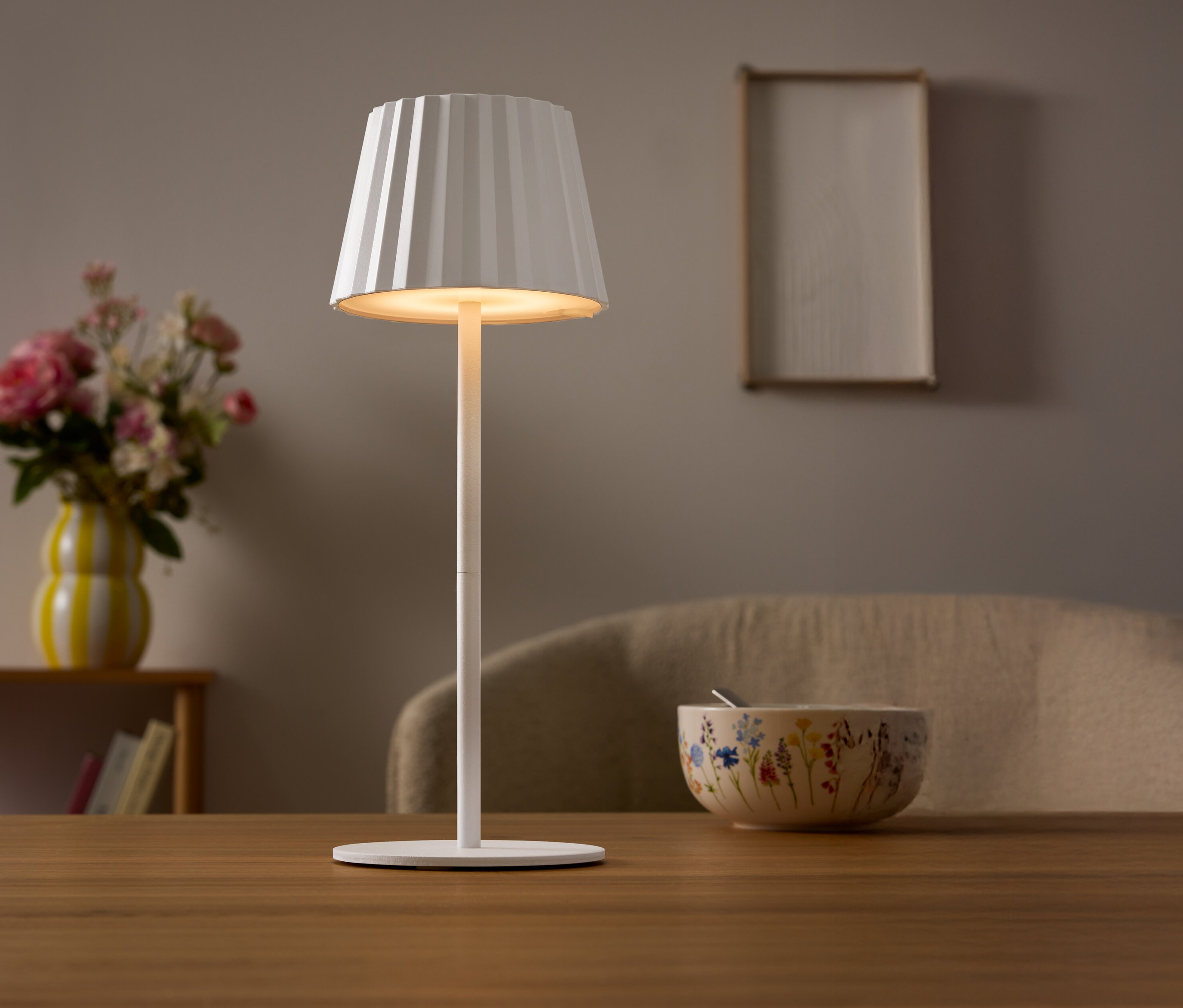 Na stole stojí biela lampa s plisovaným tienidlom. V pozadí je váza s kvetmi a miska.