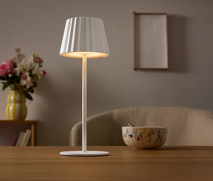 Na stole stojí biela lampa s plisovaným tienidlom. V pozadí je váza s kvetmi a miska.
