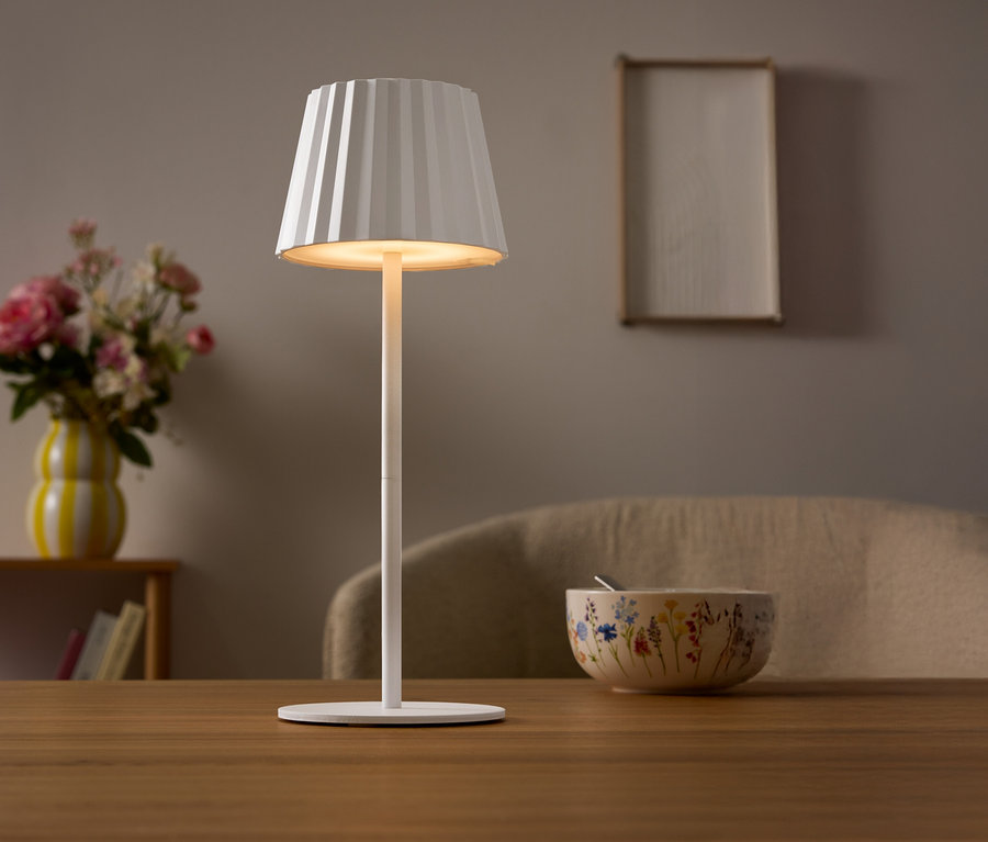 Na stole stojí biela lampa s plisovaným tienidlom. V pozadí je váza s kvetmi a miska.